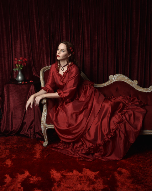  Portret ładnej modelki z rudymi włosami w efektownej historycznej wiktoriańskiej czerwonej sukni balowej. Pozowanie z humorzastym ciemnym tłem. - Zdjęcie, obraz