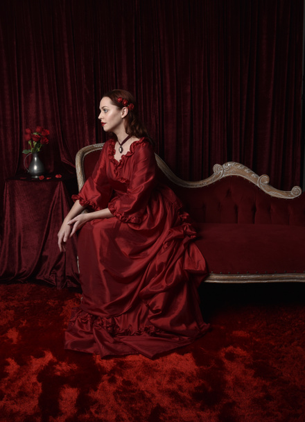  retrato de modelo femenina bonita con el pelo rojo usando glamoroso vestido de baile rojo victoriano histórico. Posando con un fondo oscuro malhumorado. - Foto, Imagen