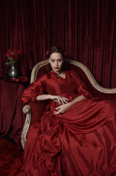  retrato de modelo femenina bonita con el pelo rojo usando glamoroso vestido de baile rojo victoriano histórico. Posando con un fondo oscuro malhumorado. - Foto, imagen
