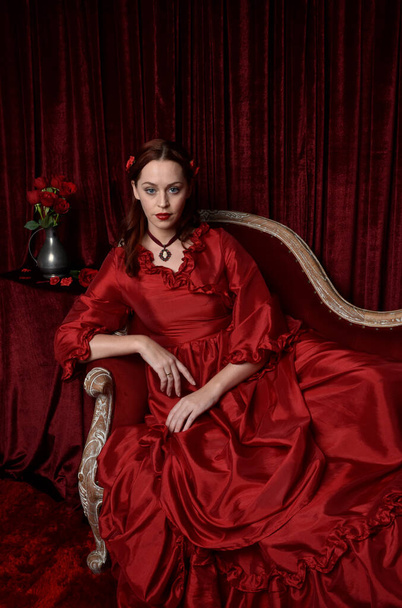  portrait of pretty female model with red hair wearing glamorous historical victorian red ballgown.  Posing with a moody dark background. - Φωτογραφία, εικόνα