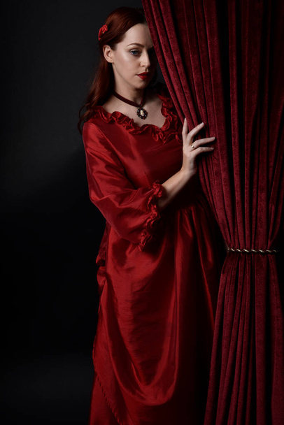  retrato de modelo femenina bonita con el pelo rojo usando glamoroso vestido de baile rojo victoriano histórico. Posando con un fondo oscuro malhumorado. - Foto, imagen