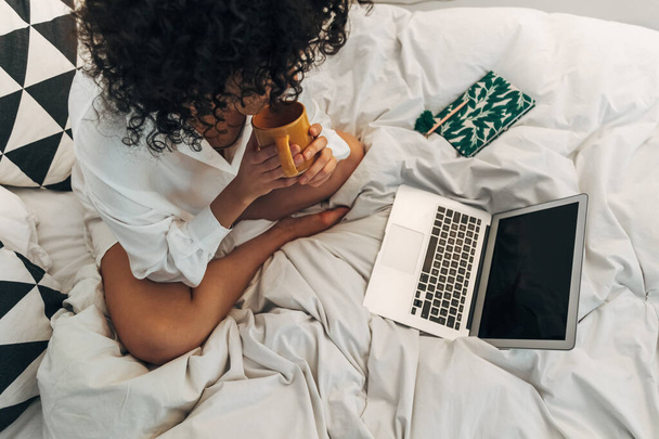 Вид сверху на молодую афроамериканку, работающую дома с ноутбуком, сидящую на кровати и пьющую чай. - Фото, изображение