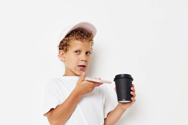 Photo portrait curly little boy in a white t-shirt cap with a phone in a glass with a drink light background unaltered - Fotografie, Obrázek