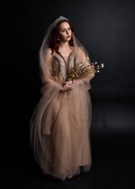  Full length portrait of pretty female model with red hair wearing glamorous fantasy tulle gown and crown.  Posing with a moody dark background. - Фото, изображение