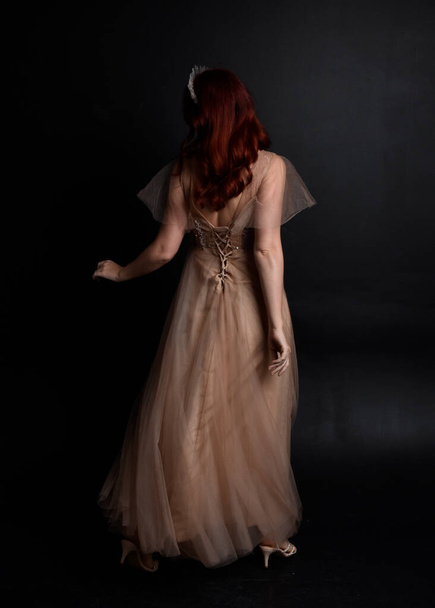 Ganzkörperporträt eines hübschen weiblichen Modells mit roten Haaren, das ein glamouröses Fantasie-Tüllkleid und eine Krone trägt. Posieren mit einem launischen dunklen Hintergrund. - Foto, Bild