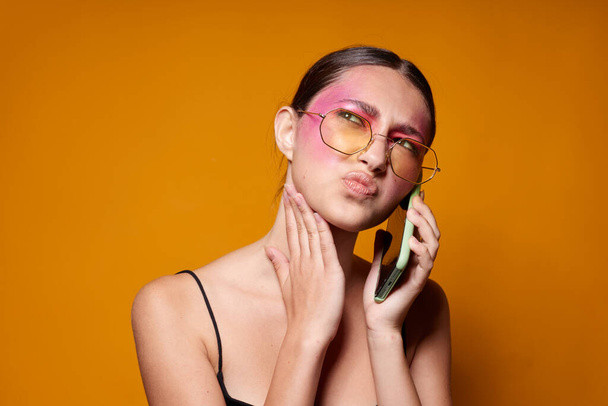 Sexy brunette woman with phone pink face makeup posing attractive look glasses yellow background unaltered - Фото, зображення