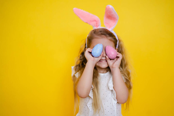 schönes lächelndes blondes Mädchen mit Hasenohren, das ein Osterei in der Hand hält, schließt die Augen, auf gelbem Hintergrund, Kind feiert Ostern. - Foto, Bild