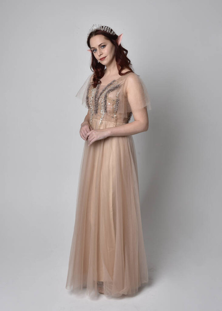 Portrait pleine longueur d'un joli modèle féminin aux cheveux roux portant une robe et une couronne en tulle fantaisie glamour. Poser avec des bras gestuels sur un fond de studio - Photo, image