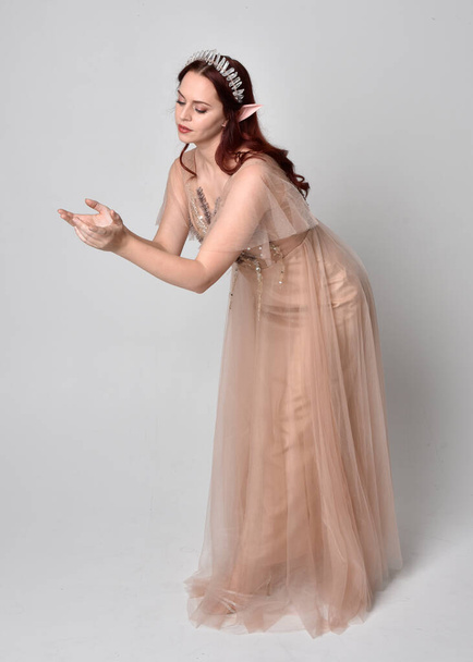 Full length portrait of pretty female model with red hair wearing glamorous fantasy tulle gown and crown.  Posing with gestural arms on a studio background - Valokuva, kuva
