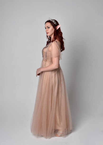 Full length portrait of pretty female model with red hair wearing glamorous fantasy tulle gown and crown.  Posing with gestural arms on a studio background - Foto, afbeelding