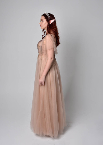 Portret pięknej modelki z rudymi włosami w efektownej sukni fantasy tiulu i koronie. Pozowanie z gestykulacyjnymi ramionami na tle studia - Zdjęcie, obraz