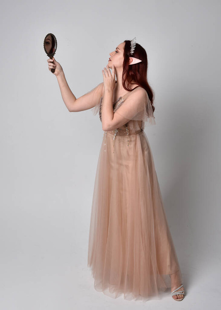 Porträt eines hübschen weiblichen Modells mit roten Haaren in glamourösem Fantasie-Tüllkleid und Krone. Posieren mit gestenreichen Armen, die einen Handspiegel auf einem Studiohintergrund halten - Foto, Bild