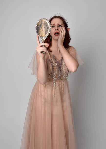 華やかなファンタジーチュールのガウンと王冠を身に着けている赤い髪のきれいな女性モデルの肖像画。スタジオの背景に手鏡を持っているジェスチャーアームでポーズをとる - 写真・画像