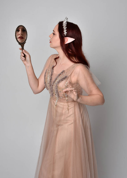 portrait of pretty female model with red hair wearing glamorous fantasy tulle gown and crown.  Posing with gestural arms  holding a hand mirror on a studio background - Photo, Image