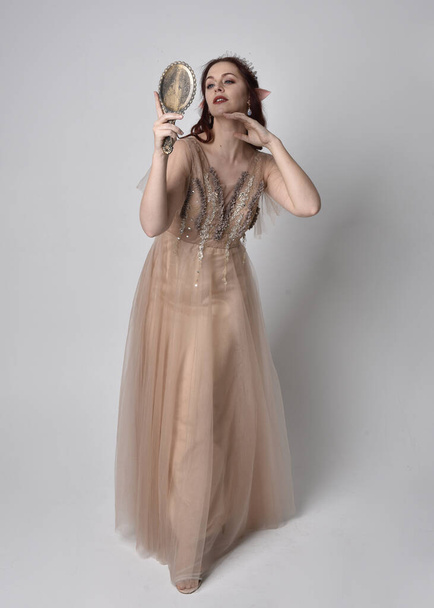 Porträt eines hübschen weiblichen Modells mit roten Haaren in glamourösem Fantasie-Tüllkleid und Krone. Posieren mit gestenreichen Armen, die einen Handspiegel auf einem Studiohintergrund halten - Foto, Bild