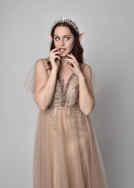 portrait of pretty female model with red hair wearing glamorous fantasy tulle gown and crown.  Posing with gestural arms on a studio background - Foto, immagini