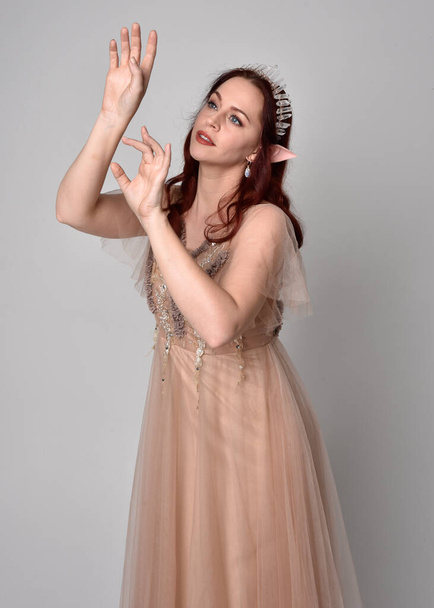 portrait of pretty female model with red hair wearing glamorous fantasy tulle gown and crown.  Posing with gestural arms on a studio background - Photo, Image