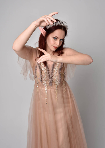 портрет красивой женщины-модели с рыжими волосами в гламурном фантазийном тюлевом платье и короне. Пэддок с жестикуляционными руками на студийном фоне - Фото, изображение