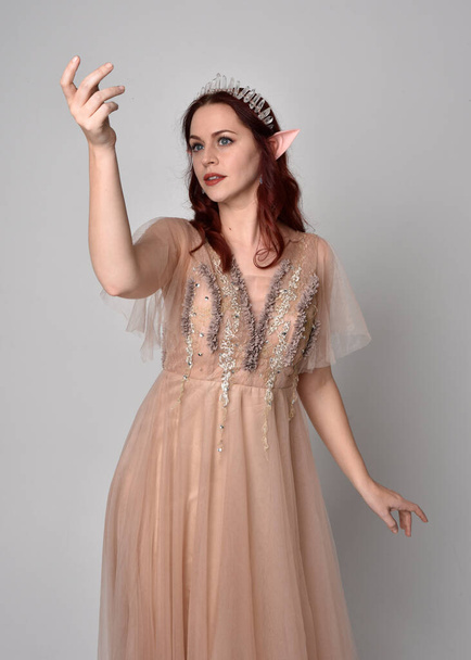 portrait of pretty female model with red hair wearing glamorous fantasy tulle gown and crown.  Posing with gestural arms on a studio background - Φωτογραφία, εικόνα