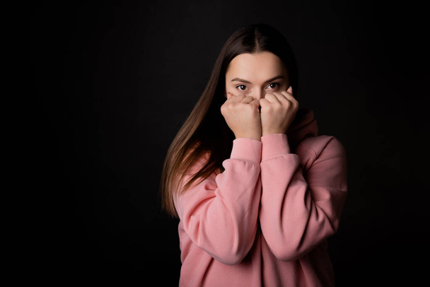 A cute young brunette in a pink hoodie hides her face with her hands, - Photo, Image
