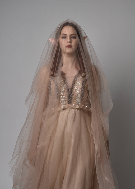  Full length portrait of pretty female model with red hair wearing glamorous fantasy tulle gown, crown and shroud veil.  Posing with gestural arms on a studio background - Photo, Image
