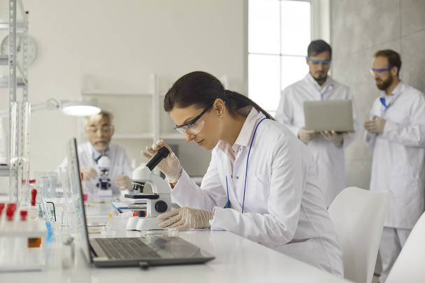 Concentrated female virologist sits at table in laboratory and examines cells under microscope. - Zdjęcie, obraz