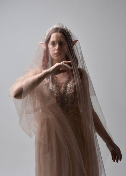 Full length portrait of pretty female model with red hair wearing glamorous fantasy tulle gown, crown and shroud veil.  Posing with gestural arms on a studio background - Photo, Image