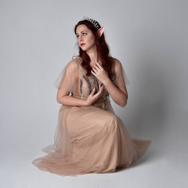 Full length portrait of pretty female model with red hair wearing glamorous fantasy tulle gown, crown and shroud veil.  Posing in a seated kneeling pose on a studio background - Photo, Image
