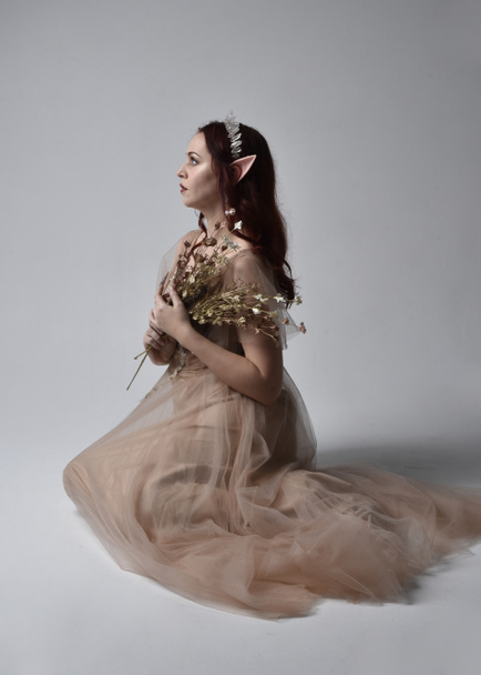 Full length portrait of pretty female model with red hair wearing glamorous fantasy tulle gown, crown and shroud veil.  Posing in a seated kneeling pose on a studio background - Photo, image