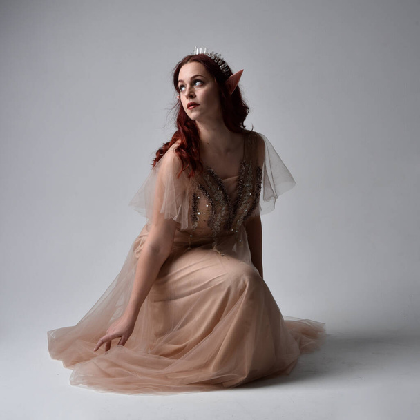 Portrait pleine longueur d'un joli modèle féminin aux cheveux roux portant une robe fantaisie glamour en tulle, une couronne et un voile enveloppant. Poser dans une pose assise à genoux sur un fond de studio - Photo, image