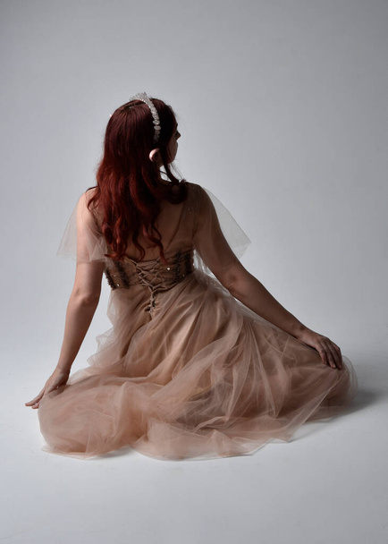 Ganzkörperporträt eines hübschen weiblichen Modells mit roten Haaren, das ein glamouröses Fantasie-Tüllkleid, Krone und Schleier trägt. Posieren in sitzender kniender Pose auf Studiohintergrund - Foto, Bild