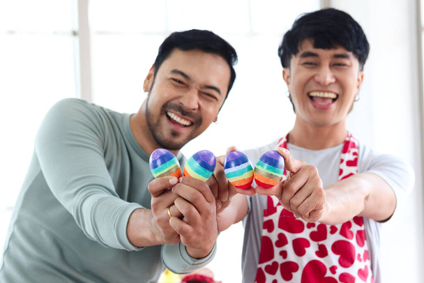 Happy young LGBT couple holding rainbow Easter eggs that are symbol of LGBTQ flag color, Asian gay male lover sharing special moment together to celebrate Easter holiday. - Photo, Image