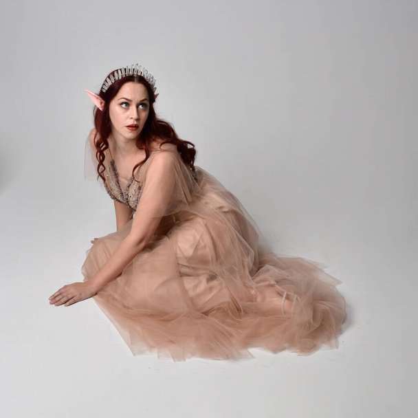 Portrait pleine longueur d'un joli modèle féminin aux cheveux roux portant une robe fantaisie glamour en tulle, une couronne et un voile enveloppant. Poser dans une pose assise à genoux sur un fond de studio - Photo, image
