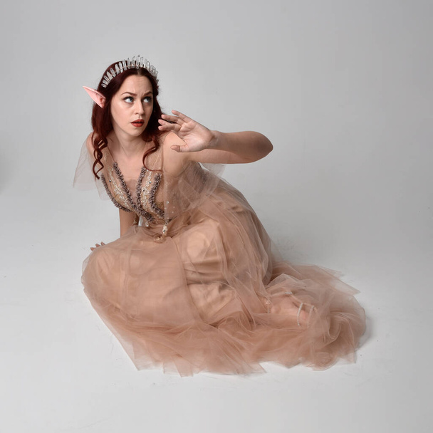 Full length portrait of pretty female model with red hair wearing glamorous fantasy tulle gown, crown and shroud veil.  Posing in a seated kneeling pose on a studio background - 写真・画像