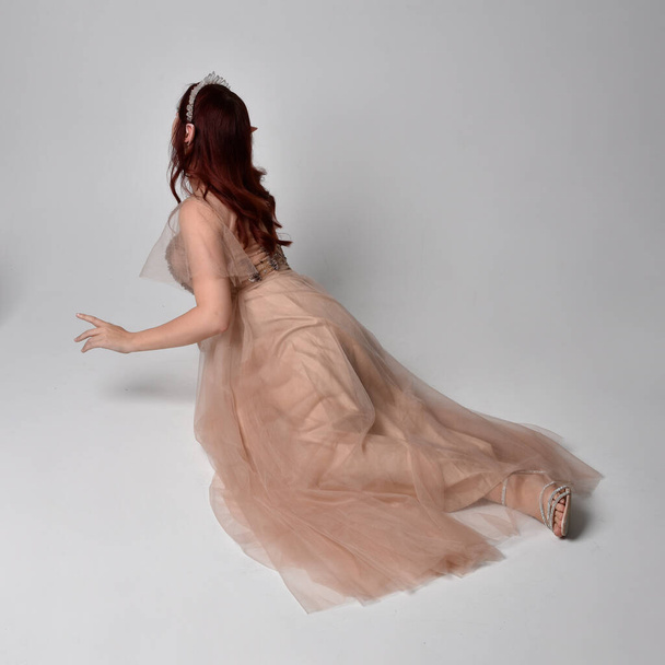 Retrato de cuerpo entero de una bonita modelo femenina de pelo rojo con glamoroso vestido de tul de fantasía, corona y velo de mortaja. Posando en una pose sentada arrodillada sobre un fondo de estudio - Foto, imagen