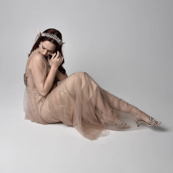 Full length πορτραίτο του όμορφου γυναικείου μοντέλου με κόκκινα μαλλιά που φορούν glamorous fantasy tulle φόρεμα, στέμμα και πέπλο. Τοποθέτηση σε καθιστή στάση γονυπετής σε φόντο στούντιο - Φωτογραφία, εικόνα