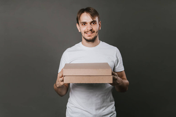 Um jovem com um sorriso em uma camiseta branca com caixas quadradas de pizza fica em um fundo cinza. Mock-up - Foto, Imagem