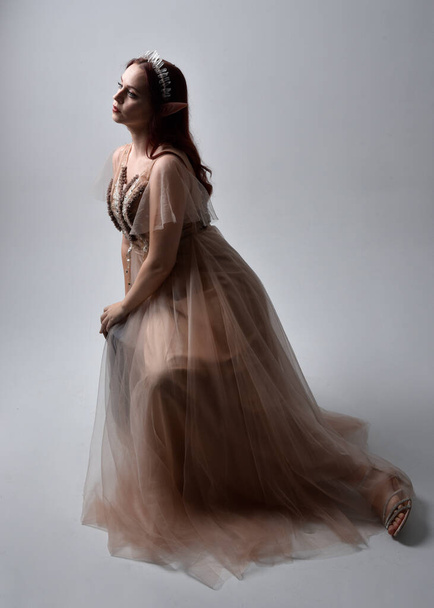  Full length portrait of pretty female model with red hair wearing glamorous fantasy tulle gown, crown. Sitting Pose with gestural arms on a studio background - Foto, imagen