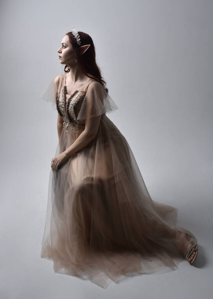  Full length portrait of pretty female model with red hair wearing glamorous fantasy tulle gown, crown. Sitting Pose with gestural arms on a studio background - Photo, Image