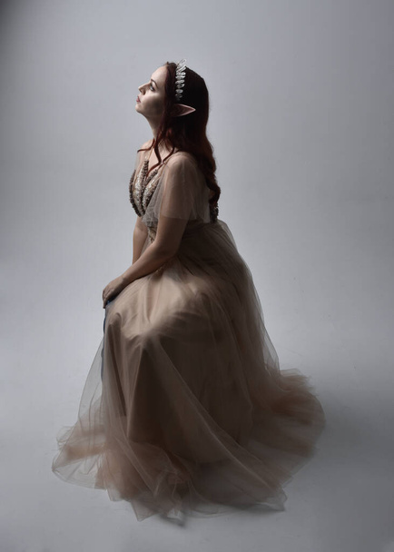  Full length portrait of pretty female model with red hair wearing glamorous fantasy tulle gown, crown. Sitting Pose with gestural arms on a studio background - Fotó, kép
