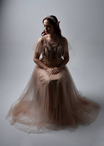  Full length portrait of pretty female model with red hair wearing glamorous fantasy tulle gown, crown. Sitting Pose with gestural arms on a studio background - Foto, afbeelding