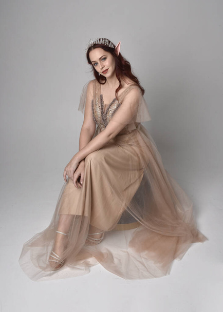  Full length portrait of pretty female model with red hair wearing glamorous fantasy tulle gown, crown. Sitting Pose with gestural arms on a studio background - Foto, Imagem