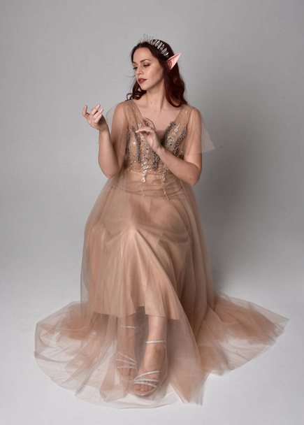  Full length portrait of pretty female model with red hair wearing glamorous fantasy tulle gown, crown. Sitting Pose with gestural arms on a studio background - Foto, imagen