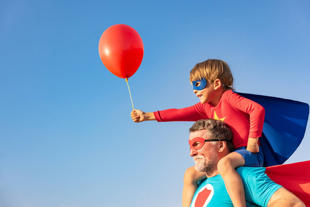 Superhero senior man and child playing outdoor. Super hero grandfather and boy having fun together against blue summer sky background. Family holiday concept. Happy Father's day - Foto, Imagem