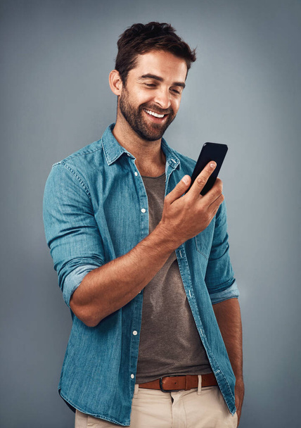The text that put a smile on his dial. Studio shot of a handsome young man using a mobile phone against a grey background. - Φωτογραφία, εικόνα