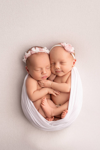 Маленькие новорожденные девочки-близнецы. Новорожденный близнец спит рядом со своей сестрой Новорожденные девочки-близнецы на фоне белого одеяла с розовыми бинтами. Девочки нежно обнимают и целуют свою сестру в милой позе. - Фото, изображение