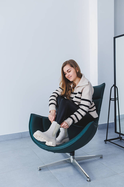 A young beautiful woman is sitting in a chair, wearing white leather boots from a new collection of shoes for spring. - Photo, Image