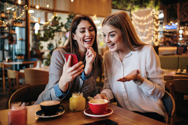 Two best friends sitting in coffee bar or restaurant after shopping and happily talking together. They are using smart phone to take selfie photo. - Fotó, kép