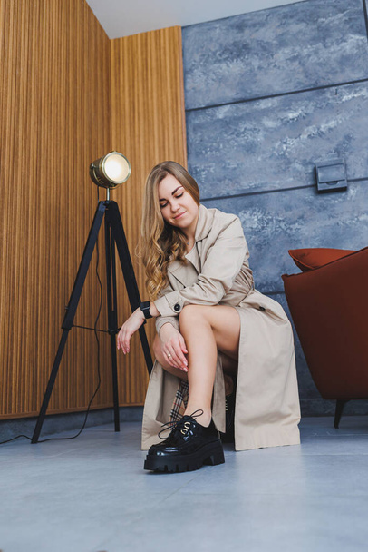 Młoda piękna kobieta siedzi na podłodze w beżowym wiosennym płaszczu przeciwdeszczowym i nowoczesnych stylowych czarnych butach. Stylowa kobieta o europejskim wyglądzie z długimi blond włosami - Zdjęcie, obraz