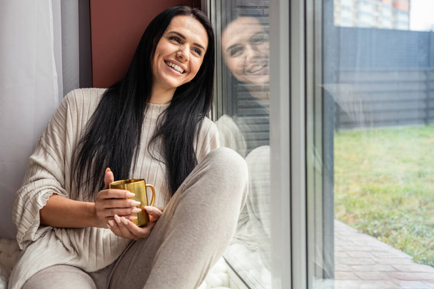 Happy beautiful brunette lady laughing enjoying weekend sitting on windowsill with tea mug relax - Foto, Imagen